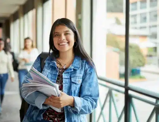 mulher de cabelos pretos lisos, usa jaqueta jeans, segura livros; ao fundo vemos pessoas e janelas abertas.