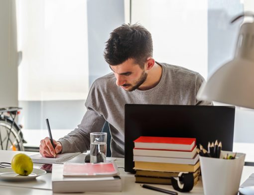 homem veste camisa cinza está olhando para caderno em que escreve; sob a mesa há notebook, livros empilhados, um copo de água e outros itens.