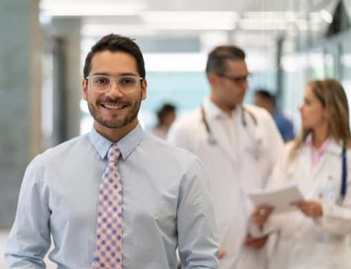 homem usa óculos transparente, camisa e gravata; no segundo plano há dois médicos conversando e o fundo é de um hospital.