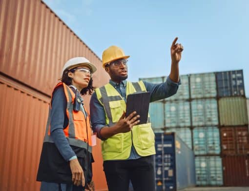 homem e mulher lado a lado usando roupas de segurança de obras; o homem aponta o alto segurando um tablet com outra mão; ao fundo vemos containers laranja e verdes.