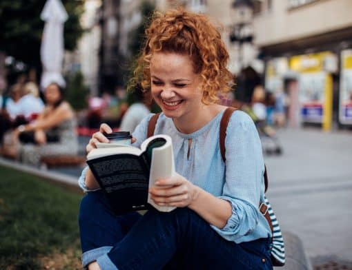 mulher branca, ruiva de cabelos cacheados, tem expressão feliz enquanto segura um livro dobrado; ela usa camisa azul e calça escura; ao fundo é possível ver uma praça com várias pessoas.