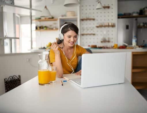 mulher branca veste blusa laranja e fones de ouvido brancos, olha sorridente para notebook branco sob a mesa também branca; ao lado vemos uma jarra e um suco de laranja e atrás dela itens de decoração de ambiente.