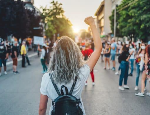 foto de mulher de costas com cabelo descolorido, mochila nas costas vestindo camiseta branca; ela tem o punho cerrado ao alto e olha várias pessoas reunidas em uma rua.