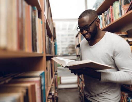 homem preto usa óculos, camisa branca e está de pé no corredor de uma livraria sorrindo e olhando um livro aberto.