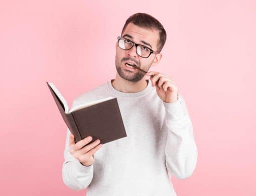 fundo rosa com homem vestindo blusa de manga comprida rosa, segura caderno marrom e morde caneta, olhando para cima.