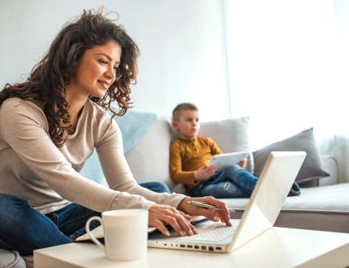 mulher morena de cabelos cacheados veste blusa de manga comprida branca e calça jeans, está digitando em um notebook branco repousado em uma mesa com uma caneca branca ao lado; no segundo plano há um menino loiro de blusa amarela e calça azul com um tablet na mão.