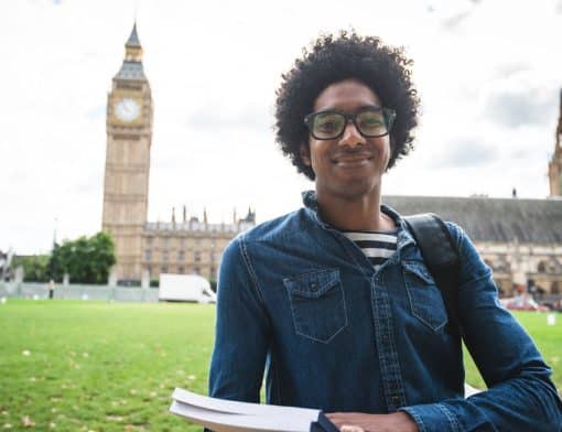 homem preto usa cabelo black power, camisa jeans e segura livros; ao fundo vemos grama e o relógio Big Ben.