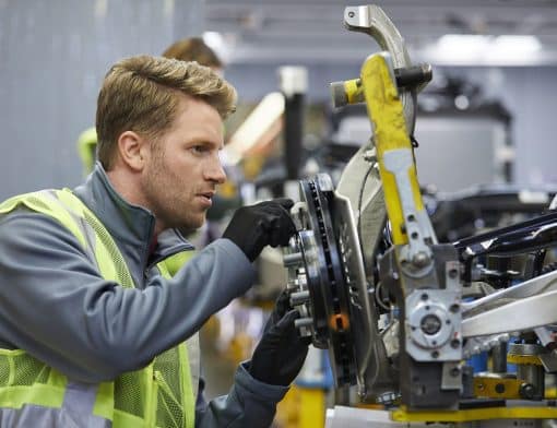 foto de uma peça de máquina industrial do lado direito e um homem loiro está montando alguma peça nela; ele usa camisa cinza de manga comprida, luvas pretas e colete de segurança.