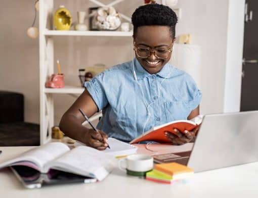 mulher preta de cabelo curto, usa óculos e camisa azul de manga curta; ela segura livro de capa vermelha e escreve em cadernos abertos sob a mesa, que tem notebook, post it e outros itens. Ao fundo vemos estante branco com decorações.