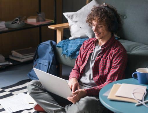 homem de cabelo cacheado, veste camisa vermelha e calça cinza, está sentado no chão com um notebook no colo; atrás dele há um sofá com almofada, ao seu lado há uma estante de um lado e uma mesa azul baixa com papeis de outro.