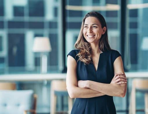 imagem de mulher branca sorridente de cabelos lisos claro, usa roupa azul escuro e tem os braços cruzados; ao fundo é possível ver janelas de vidro, cadeiras e abajur.