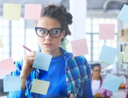 mulher de cabelo preso usa camiseta azul e camisa xadrez e óculos, ela olha vidro com vários post its colados enquanto escreve em um.