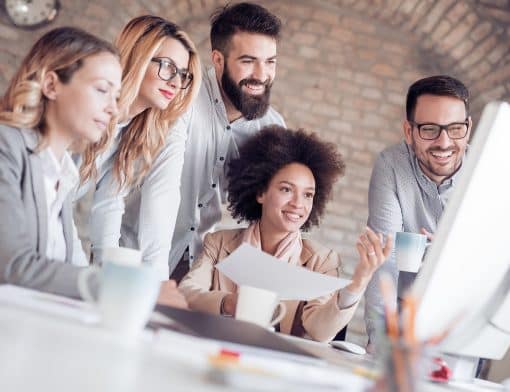 ao redor de uma mesa branca há uma pessoa preta de cabelo black power, segura uma folha, e do lado dela há dois homens e duas mulheres olhando o monitor sob a mesa, que tem notebook, caneca e outros papeis.