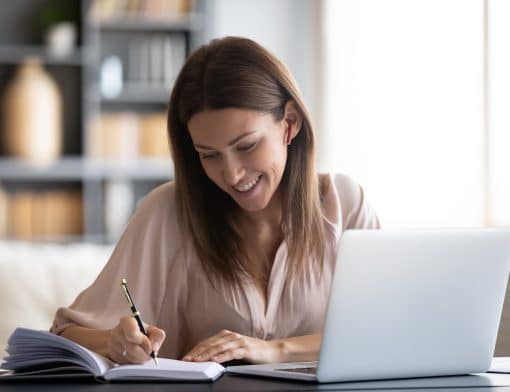 mulher branca de cabelos lisos, usa blusa rosa claro, ela está apoiada em uma mesa escrevendo em um caderno com um notebook branco ao lado; no fundo vemos uma estante escura e parede branca.