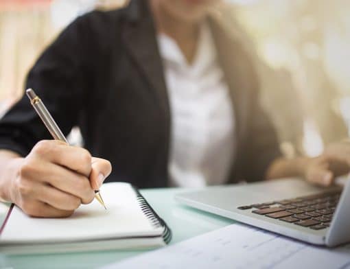 foco no busto de uma mulher de camisa branca e brazer preto, ela escreve em um caderno e com outra mão digita no notebook.