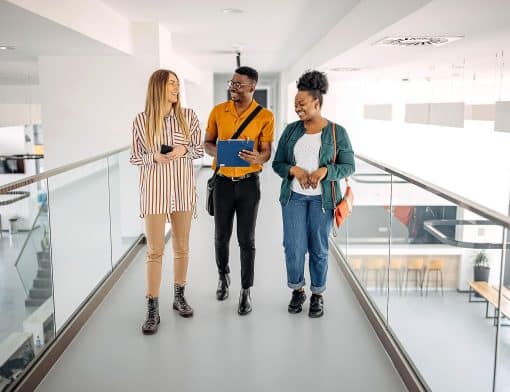 três pessoas aparecem na foto: uma mulher de cabelo enrolado, um homem negro e uma mulher loira. Todos parecem felizes ao andar por uma passarela. A primeira usa uma camisa listrada e segura o celular; o homem, usando uma camisa laranja e com uma prancheta em mãos; e a terceira com uma bolsa a tira colo e jaqueta verde oliva.