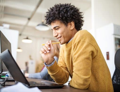 homem preto de cabelo cacheado usa blusa amarela, tem os braços apoiados na mesa, enquanto olha um notebook sob a mesa; ao fundo vemos luzes e outras pessoas apoiadas na mesa.