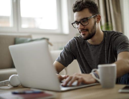 imagem de homem branco, de cabelos pretos, usa óculos e veste camiseta cinza; ele está digitando em um notebook cinza repousado em uma mesa; ao fundo vemos cenário de uma casa.