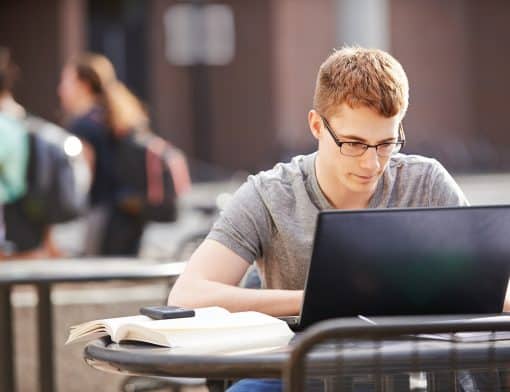 homem branco e loiro está sentado ao ar livre mexendo em seu notebook preto; ele usa óculos e camiseta cinza; ao seu lado é possível ver livros e um celular; ao fundo pessoas e muros de uma escola.