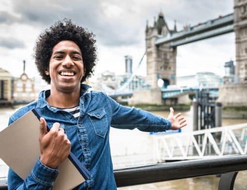 jovem preto de cabelo afro está sorrindo, veste um casaco jeans e segura alguns papeis com a mão direita enquanto aponta com a esquerda a Tower Bridge de Londres ao fundo.