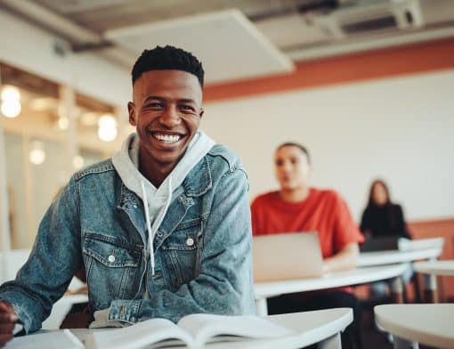 homem jovem preto sorri para a foto, usa jaqueta jeans e apoia sob uma mesa com livro; ao fundo vemos outros estudantes.