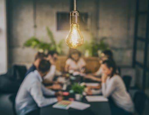 lâmpada acesa no primeiro plano na parte superior e ao fundo pessoas diversas ao redor de uma mesa conversando e mexendo em papeis.