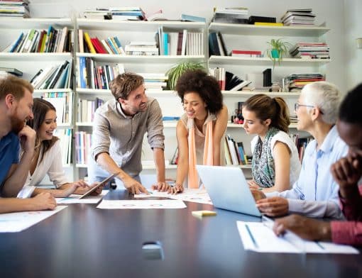 uma mesa preta toma boa parte do espaço da foto e sob ela existem papeis e notebooks; 7 pessoas estão ao redor, algumas sentadas e outras de pé inclinadas a ela; as pessoas são diversas entre homens e mulheres, de idades e etnias diferentes.