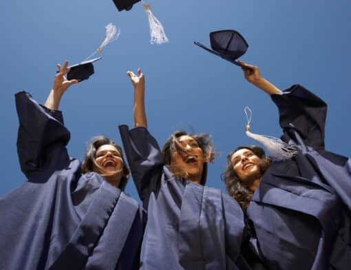 foco de baixo para cima de três mulheres com roupas de formatura, elas estão alegres com os braços para cima jogando o chapeu de formatura.