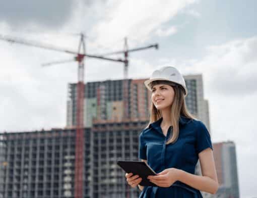 mulher usa blusa azul, tablet preto e chapeu de construção branco, olha para o horizonte e ao fundo vemos prédios e guindastes.