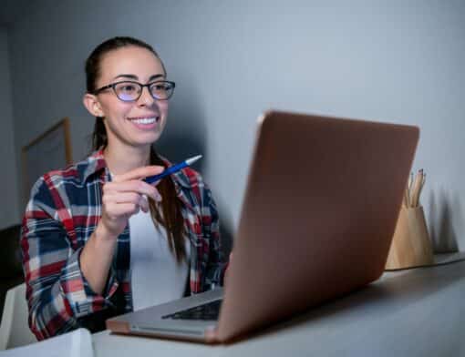 mulher tem cabelos presos usa óculos, camiseta branca e camisa xadrez, segura uma caneta no alto e olha um notebook sob uma mesa cinza.