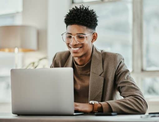 imagem de homem preto que usa camisa marrom e blazer de mesma cor, ele está sorrindo olhando para um notebook.