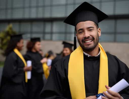 homem está com roupa e chapéu de formatura pretos, com detalhe em amarelo, ele sorri enquanto segura o diploma; ao fundo outros colegas da mesma forma conversam atrás dele.