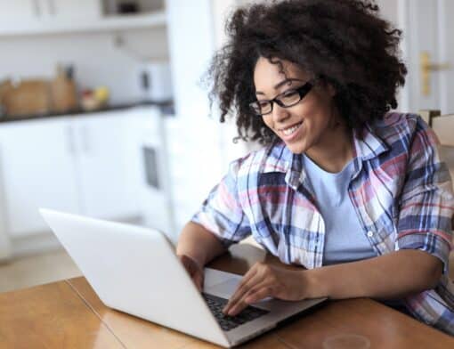 mulher preta de cabelo cacheado, usa blusa branca e camisa xadrez por cima, e óculos preto. Ela digita no notebook cinza que está em cima da mesa; ao redor é possível ver itens de cozinha.