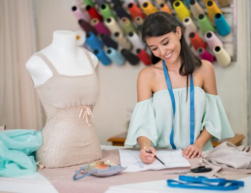 mulher de cabelos castanhos, usa blusa azul, tem fita métrica ao redor do pescoço, escreve em um papel; em cima da mesa vemos tecidos, papeis e um manequim vestido; ao fundo, rolos de linha coloridos.