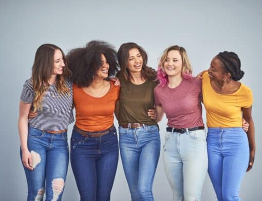 imagem de fundo cinza com 5 mulheres abraçadas diversas vestidas com camisetas coloridas e calças jeans;