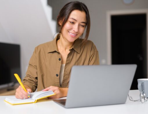 mulher branca de cabelo preso, veste camisa caramelo, está sorrindo olhando para um notebook sob uma mesa, enquanto escreve em um caderno; ao fundo vemos uma porta e uma tv.