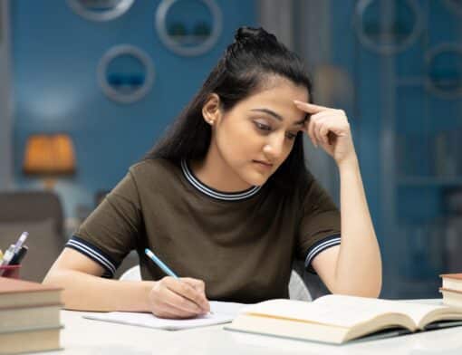 foco de mulher de cabelos pretos, veste camiseta marrom escuro, sentada na frente de uma mesa escrevendo em um papel enquanto olha para um livro aberto; ela tem uma mão na testa e vemos livros empilhados na mesa; no fundo vemos parede azul com decorações.