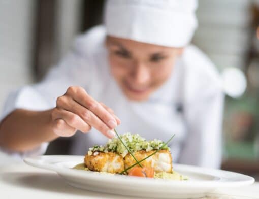 foto de uma chef decorando um prato de comida; sua mão e o prato estão no primeiro plano e seu rosto e corpo embaçados no segundo plano ao fundo.