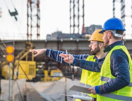 dois homens vestidos com equipamentos de segurança estão lado a lado apontando algo; atrás deles é possível ver uma construção em andamento.