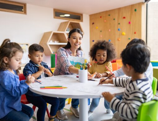 imagem de uma sala de aula infantil onde no primeiro plano tem uma professora ao centro e várias crianças diferentes do lado dela ao redor de uma mesa, pintando papeis e mexendo em lápis.