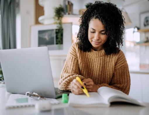 mulher preta de cabelo cacheado preto, usa sueter caramelo, grifa um livro sob a mesa; ao lado vemos um notebook e itens de estudo na mesa; ao fundo vemos itens de decoração de uma sala.