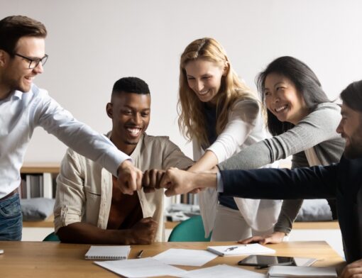 ao redor de uma mesa cheia de papeis, várias pessoas diferentes, brancas e pretas, homens e mulheres estão de pé inclinados ao tocar os pulsos todos juntos em sinal de união.