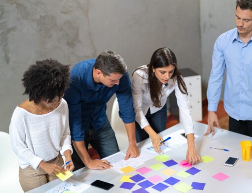 sob uma mesa branca há vários post its coloridos, canetas. Uma mulher preta de cabelo crespo, blusa branca e calça bege está do lado esquero segurando um post it, um homem de camisa azul ao seu lado, uma mulher de cabelo escuro e camisa branca ao lado e um homem de camisa azul clara de pé.