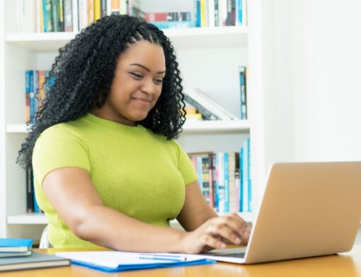 mulher preta de cabelo cacheado veste camiseta verde e mexe em notebook sob uma mesa que tem outros papeis e canetas. Ao fundo vemos estante com livros.