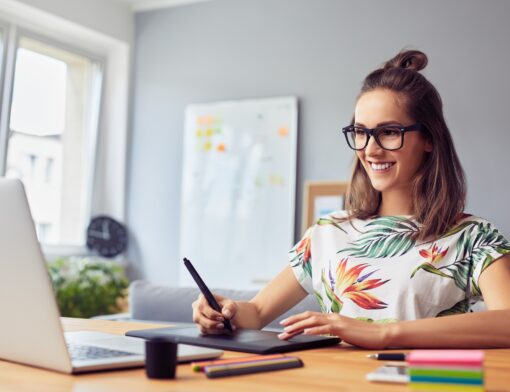 mulher usa óculos e blusa colorida, cabelo solto com coque alto; sob a mesa ela mexe em uma mesa digitalizadora enquanto olha para um notebook; ao lado há canetas, posts its e outros itens.