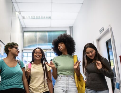 quatro mulheres estudantes de estilos diferentes estão lado a lado em um corredor escolar, elas se olham e sorriem.